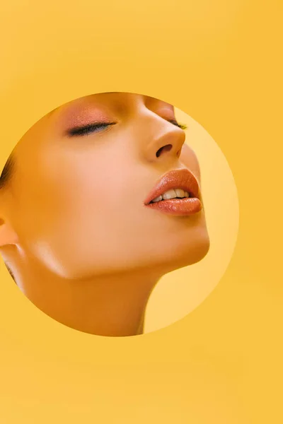 Portrait of beautiful woman with shiny makeup in paper round hole with closed eyes isolated on yellow — Stock Photo