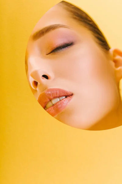 Portrait of beautiful woman with shiny makeup in paper round hole with closed eyes isolated on yellow — Stock Photo