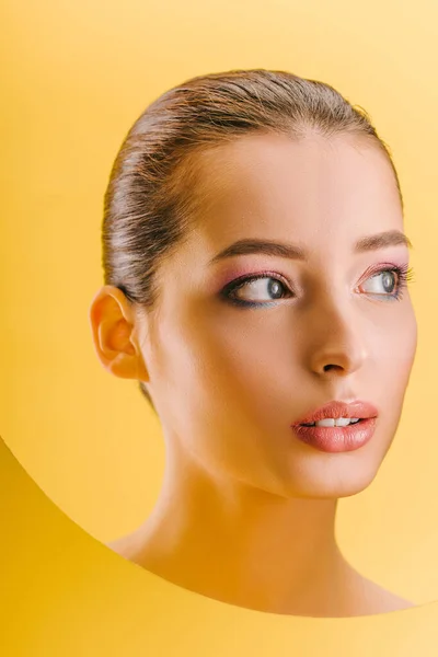 Portrait of beautiful woman with shiny makeup in paper round hole looking away isolated on yellow — Stock Photo