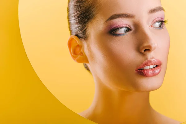 Portrait of beautiful woman with shiny makeup in paper round hole looking away isolated on yellow — Stock Photo