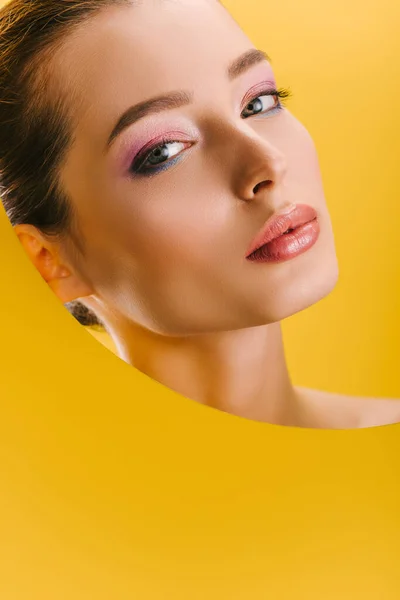 Portrait de belle femme avec un maquillage brillant dans un trou rond en papier regardant la caméra isolée sur jaune — Photo de stock