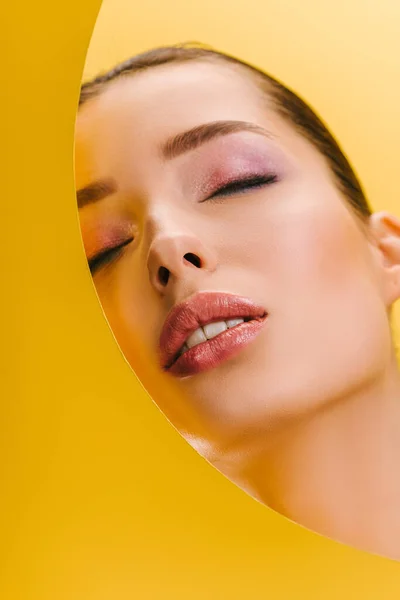 Portrait of beautiful woman with shiny makeup in paper round hole with closed eyes isolated on yellow — Stock Photo