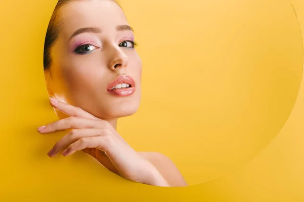 Portrait of beautiful naked woman with shiny makeup in paper round hole with hand isolated on yellow — Stock Photo