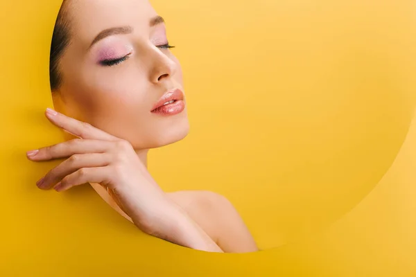 Retrato de hermosa mujer desnuda con maquillaje brillante en papel agujero redondo con la mano y los ojos cerrados aislados en amarillo - foto de stock