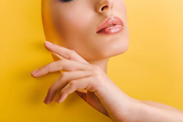 Обрізаний вид красивої жінки з блискучими губами в паперовому круглому отворі з рукою ізольовано на жовтому — стокове фото
