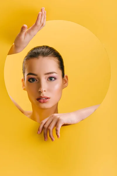 Портрет красивої жінки з блискучим макіяжем в паперовому круглому отворі з руками ізольовані на жовтому — стокове фото