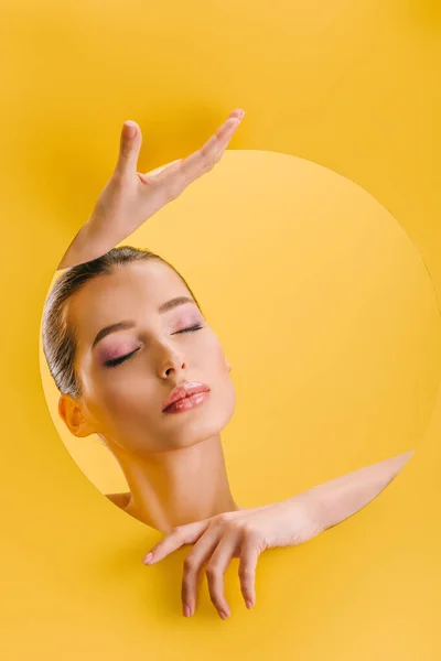 Retrato de mulher bonita com maquiagem brilhante em papel buraco redondo com as mãos e olhos fechados isolados no amarelo — Fotografia de Stock