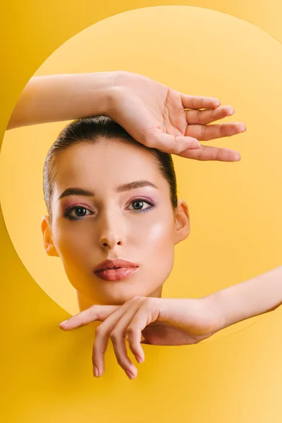 Portrait of beautiful woman with shiny makeup in paper round hole with hands isolated on yellow — Stock Photo