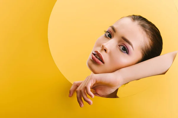 Portrait of beautiful woman with shiny makeup in paper round hole with hand isolated on yellow — Stock Photo