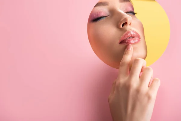Retrato de mujer hermosa con maquillaje brillante en papel rosa agujero redondo con la mano y los ojos cerrados aislados en amarillo - foto de stock