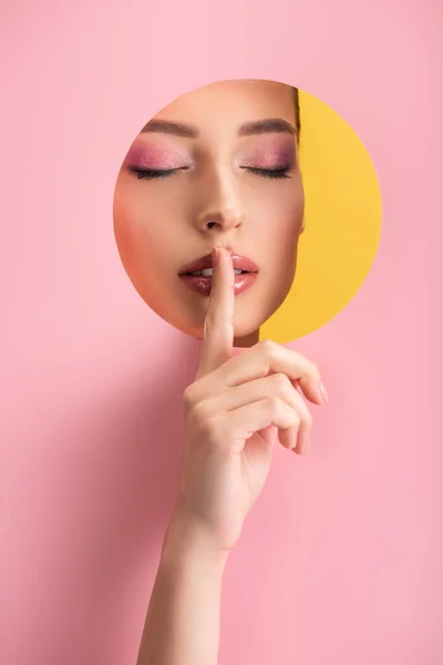 Portrait of beautiful woman with shiny makeup and closed eyes in pink paper round hole showing shh sign isolated on yellow — Stock Photo