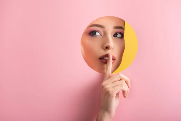 Retrato de mujer hermosa con maquillaje brillante en papel rosa agujero redondo que muestra el signo shh aislado en amarillo - foto de stock