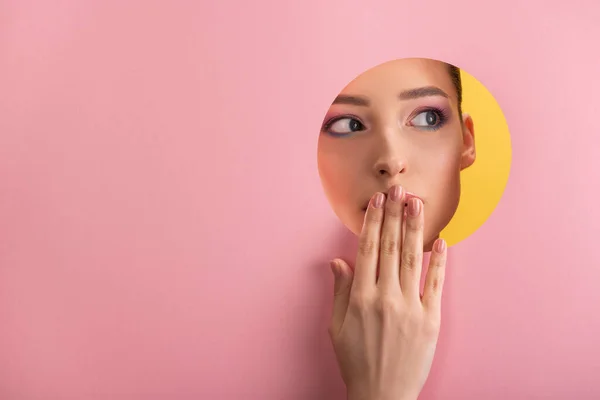 Retrato de mujer hermosa con maquillaje brillante en papel rosa agujero redondo que cubre la boca con la mano aislada en amarillo - foto de stock