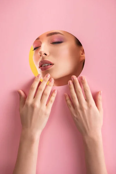 Retrato de mujer hermosa con maquillaje brillante y ojos cerrados en papel rosa agujero redondo con las manos aisladas en amarillo - foto de stock