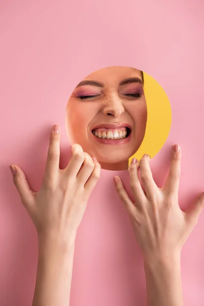 Portrait de belle femme avec maquillage brillant en papier rose trou rond montrant des dents isolées sur jaune — Photo de stock