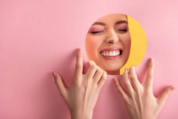 Portrait de belle femme avec maquillage brillant en papier rose trou rond montrant des dents isolées sur jaune — Photo de stock