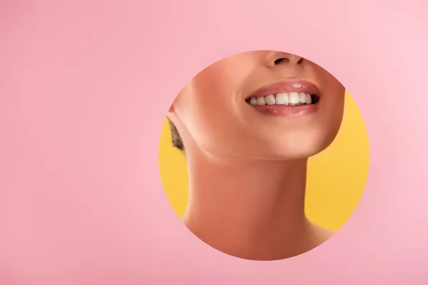 Vista parcial de hermosa mujer con labios brillantes en papel rosa agujero redondo sonriendo aislado en amarillo - foto de stock