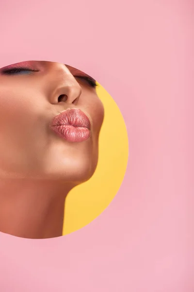 Partial view of beautiful woman with shiny lips in pink paper round hole pouting lips isolated on yellow — Stock Photo