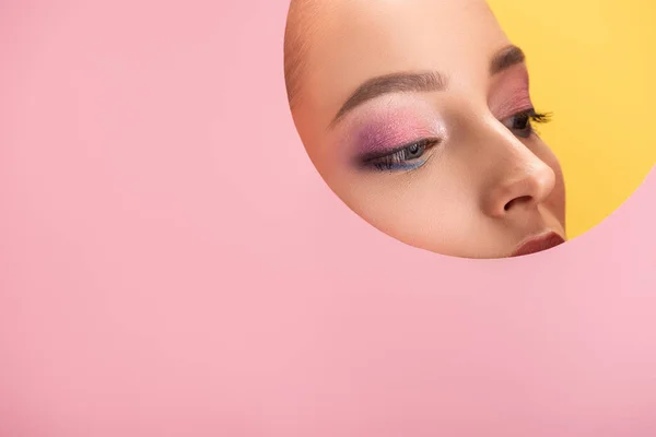 Portrait of beautiful woman with shiny eyeshadow in pink paper round hole looking away isolated on yellow — Stock Photo