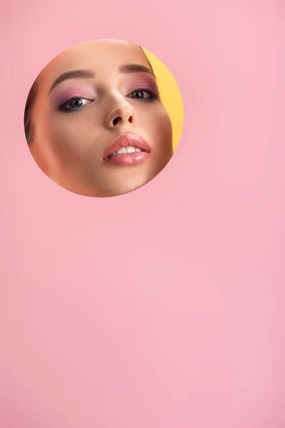 Retrato de mujer hermosa con maquillaje brillante en papel rosa agujero redondo aislado en amarillo - foto de stock