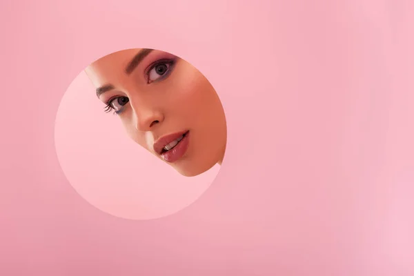 Portrait of beautiful woman with shiny makeup in paper round hole isolated on pink — Stock Photo