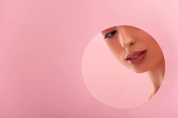 Portrait of beautiful woman with shiny makeup in paper round hole isolated on pink — Stock Photo