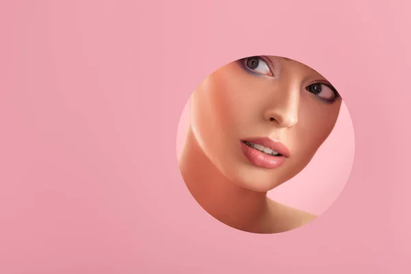 Portrait of beautiful woman with shiny makeup in paper round hole isolated on pink — Stock Photo