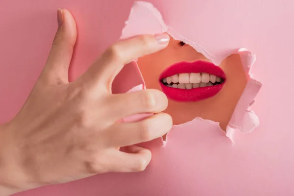 Vista recortada de hermosa mujer con labios carmesí y dientes blancos en agujero roto de papel aislado en rosa - foto de stock