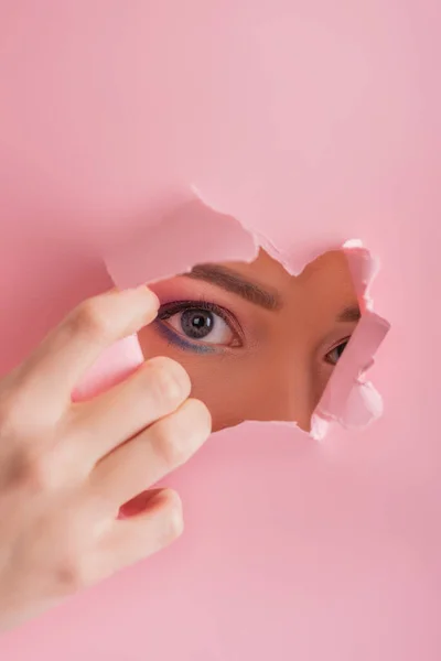 Schöne Frau mit glänzendem Make-up, die durch ein zerrissenes Loch auf rosa Papier blickt — Stockfoto