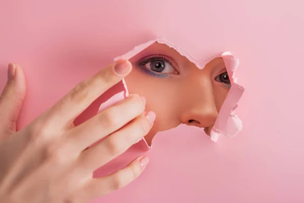 Hermosa mujer con maquillaje brillante mirando a través de agujero roto de papel aislado en rosa - foto de stock