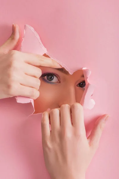 Hermosa mujer con maquillaje brillante mirando a través de agujero roto de papel aislado en rosa - foto de stock