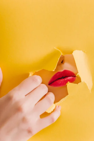 Vista recortada de hermosa mujer con labios carmesí en el agujero roto de papel amarillo - foto de stock