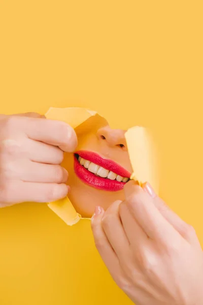 Vista recortada de la hermosa mujer con labios carmesí sonriendo en el agujero roto de papel amarillo - foto de stock