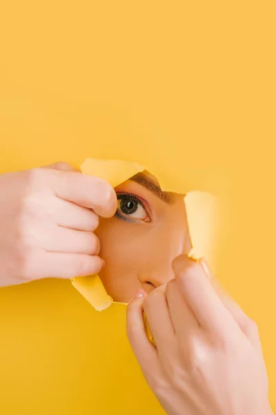 Cropped view of beautiful woman looking through yellow paper torn hole — Stock Photo