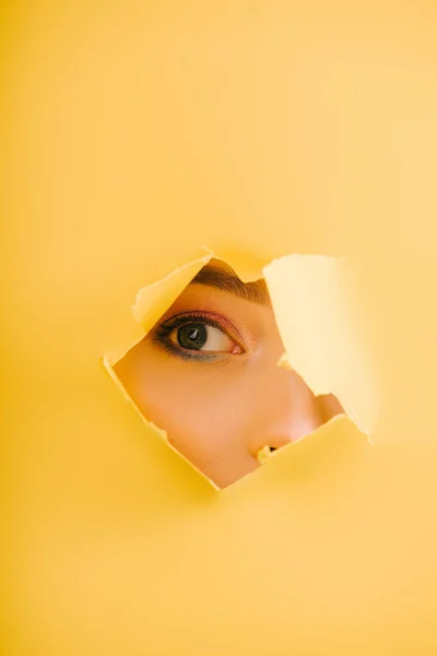 Vista recortada de la hermosa mujer mirando a través del agujero roto de papel amarillo - foto de stock