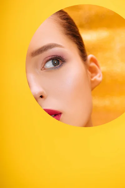 Hermosa mujer con maquillaje colorido mirando a través de agujero círculo de papel amarillo - foto de stock