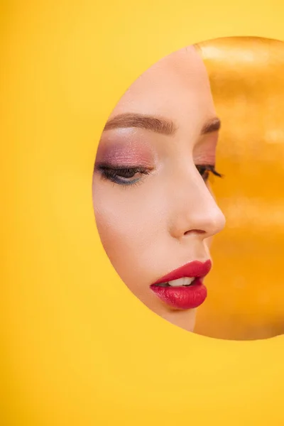 Hermosa mujer con maquillaje colorido mirando a través de agujero círculo de papel amarillo - foto de stock