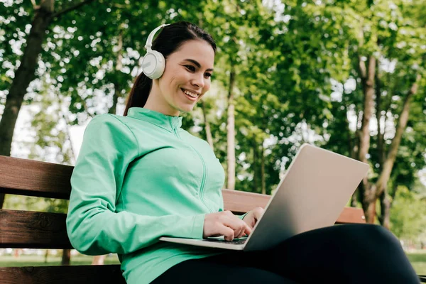 Souriante fille dans les écouteurs à l'aide d'un ordinateur portable sur le banc dans le parc — Photo de stock