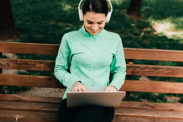 Belle freelance souriante dans les écouteurs en utilisant un ordinateur portable dans le parc — Photo de stock