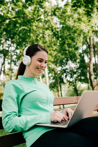 Freelancer sonriente escuchando música en auriculares y usando laptop en el parque - foto de stock