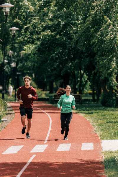 Mujer sonriente corriendo cerca de novio guapo en pista de atletismo en el parque - foto de stock