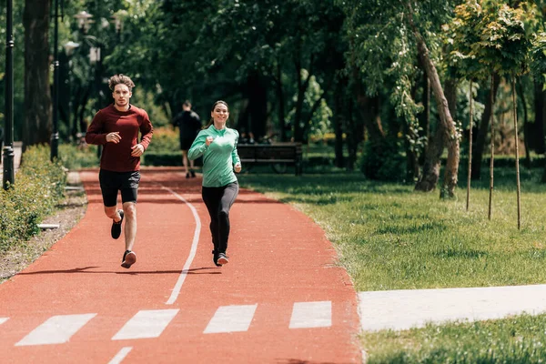 Donna sorridente in esecuzione vicino bel ragazzo sulla pista di corsa nel parco — Foto stock