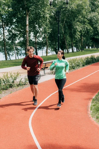 Hermosa mujer sonriendo mientras trota cerca de novio en pista de atletismo en el parque - foto de stock