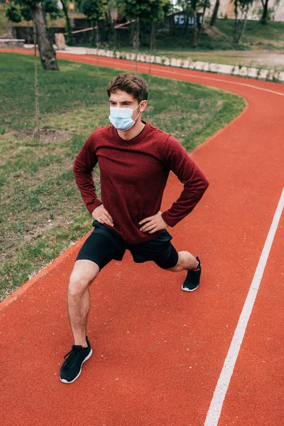 Man in medical mask doing lunges while working out on running track in park — Stock Photo