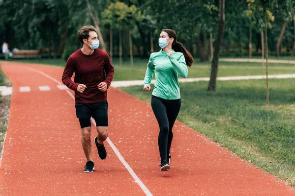 Runners in medical masks looking at each other while training on running track in park — Stock Photo