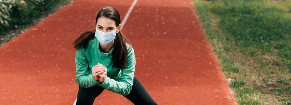 Panoramic shot of sportswoman in medical mask doing squat on running track in park — Stock Photo