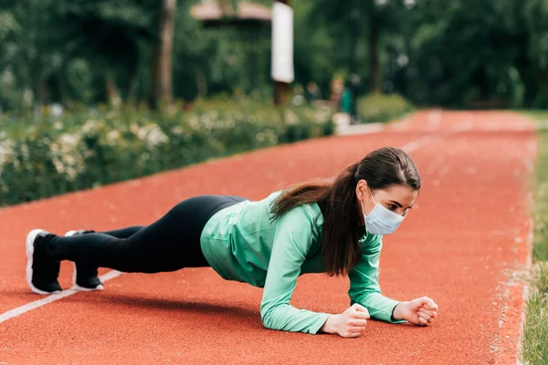 Vista lateral de la deportista en máscara médica haciendo tablón mientras hace ejercicio en el parque - foto de stock
