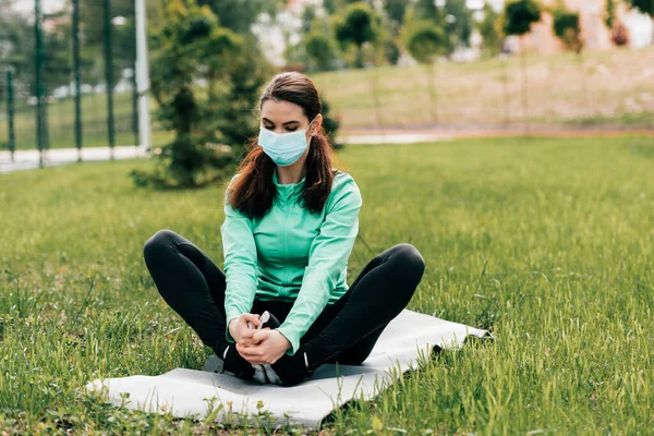 Deportiva en máscara médica haciendo ejercicio en la alfombra de fitness en la hierba en el parque - foto de stock