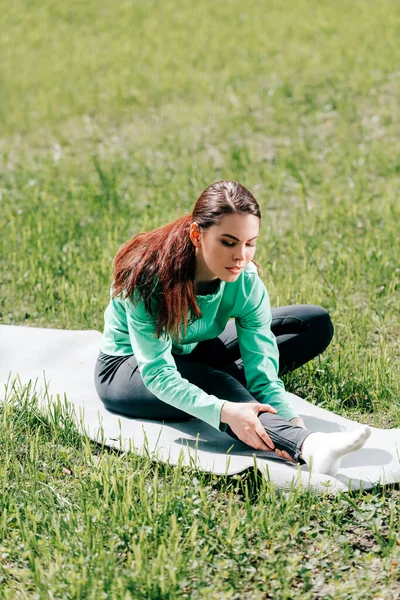 Foco seletivo de esportista atraente esticando no tapete de fitness na grama no parque — Fotografia de Stock