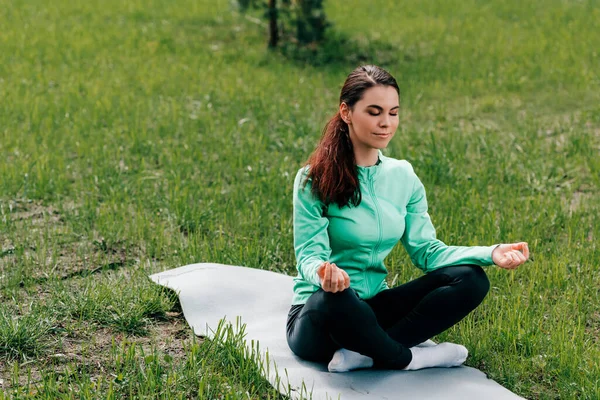 Hermosa mujer con las piernas cruzadas meditando en la alfombra de fitness en la hierba en el parque - foto de stock